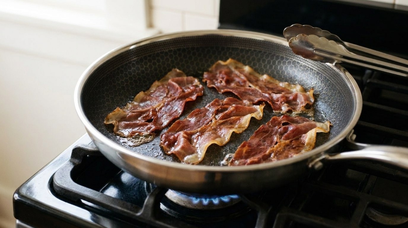 Four thin slices of Parma ham sizzling and crinkling in a dry nonstick skillet on a gas burner, darkened to deep ruby with crisp golden fat edges, before the English muffins go in